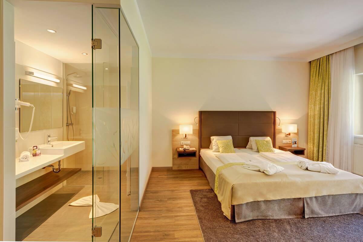 Habitación de hotel con ducha envuelta en vidrio, una cama grande con cabecera, lámparas de noche y mesita de noche.