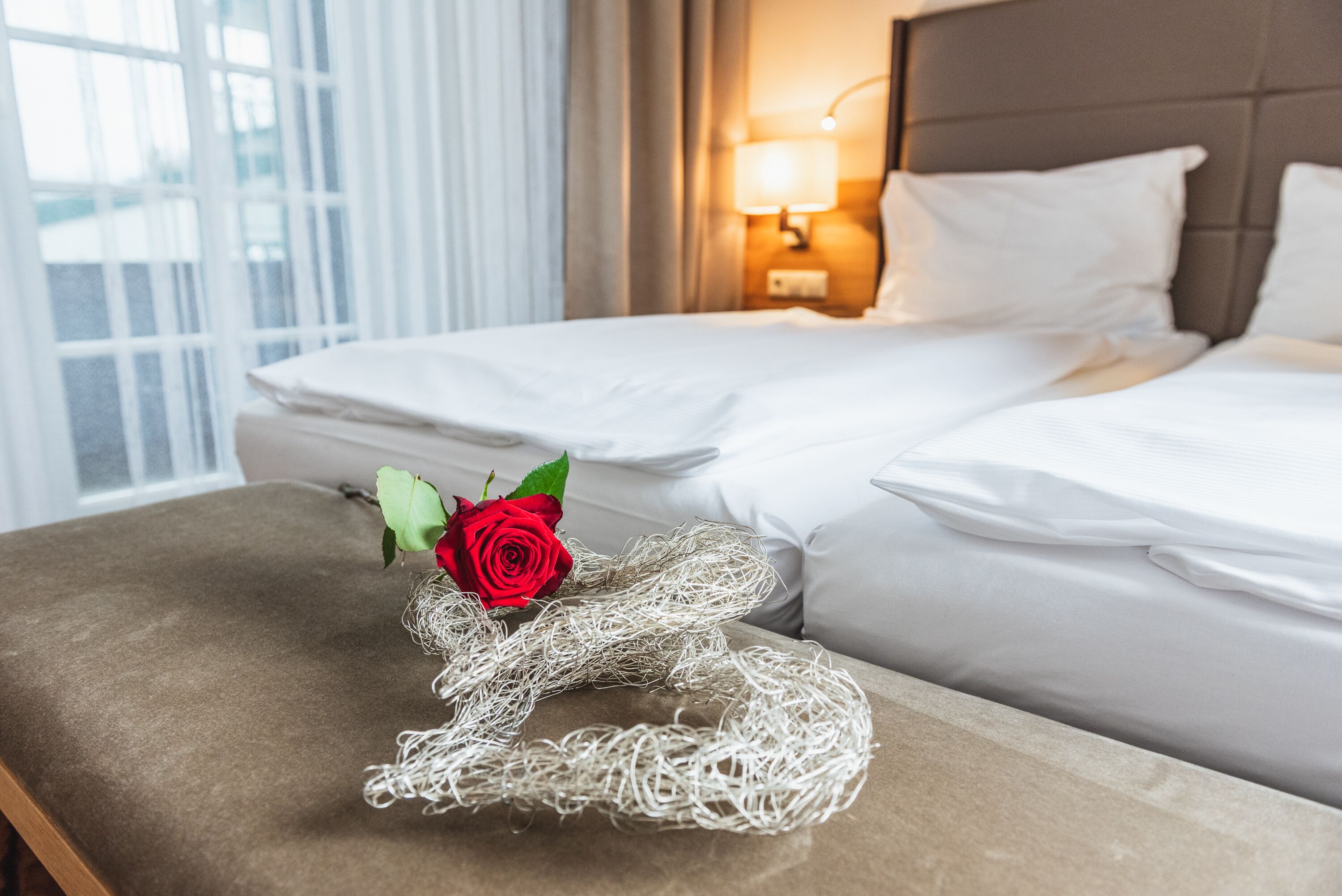 Una cama bien hecha con sábanas blancas, una lámpara de noche y una ventana con cortinas.