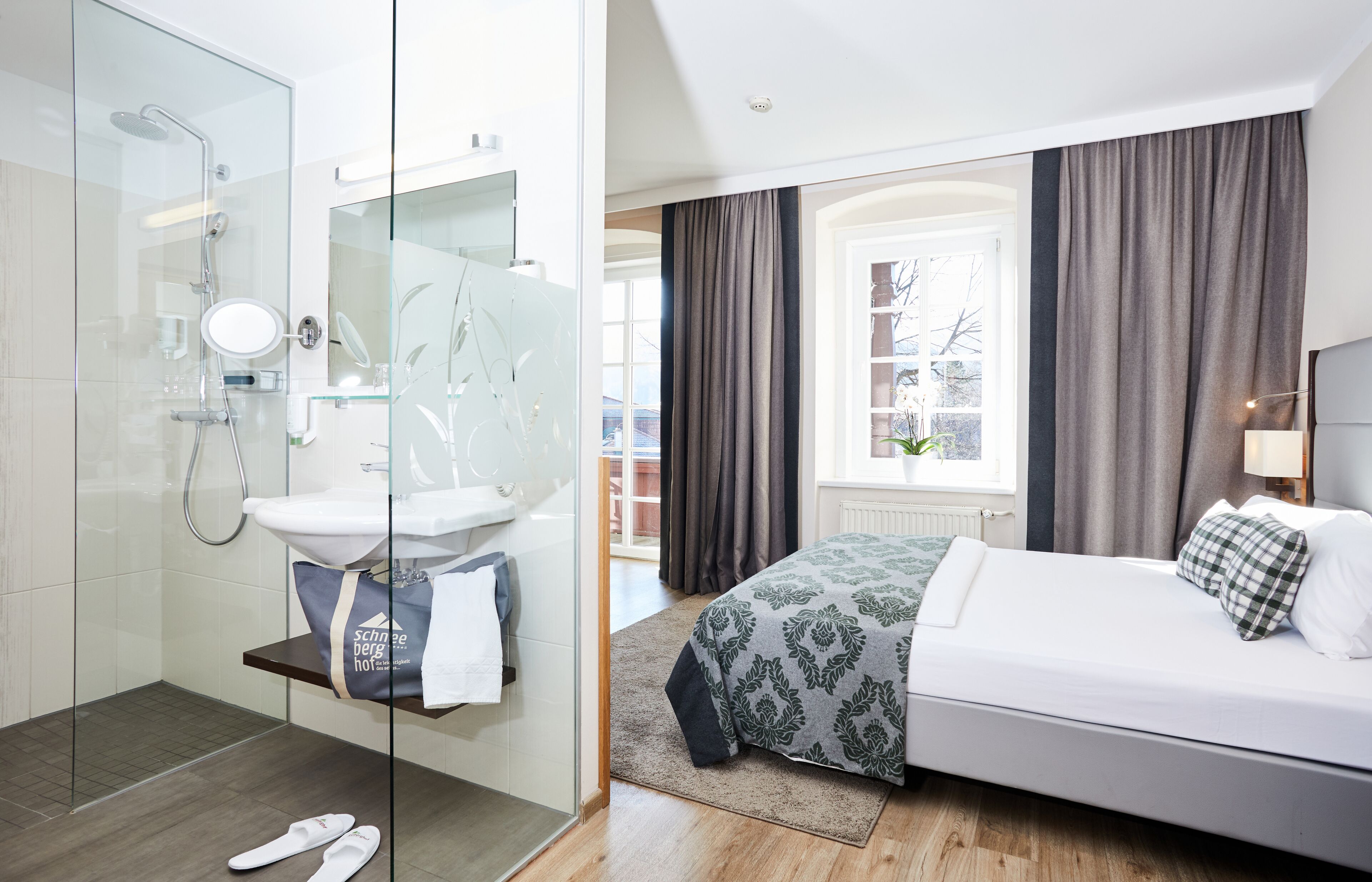 A modern hotel room with a glass shower enclosure, a white sink, a bed with a floral bedspread, and a view of the outside through large windows.