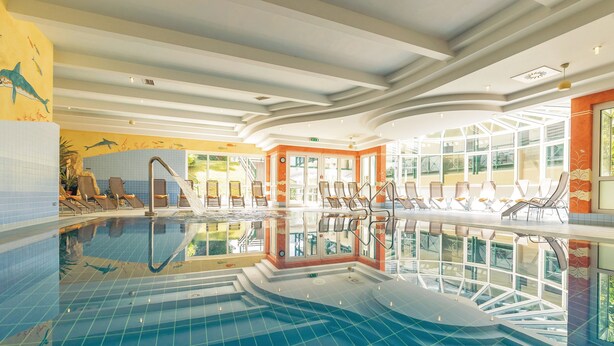 An indoor swimming pool with clear blue water, sun loungers, and a view of the outside through large windows. - Schneeberghof (Puchberg Am Schneeberg)