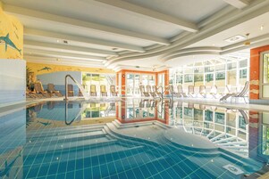Uma piscina interna com água azul cristalina, espreguiçadeiras e vista para o exterior através de grandes janelas.