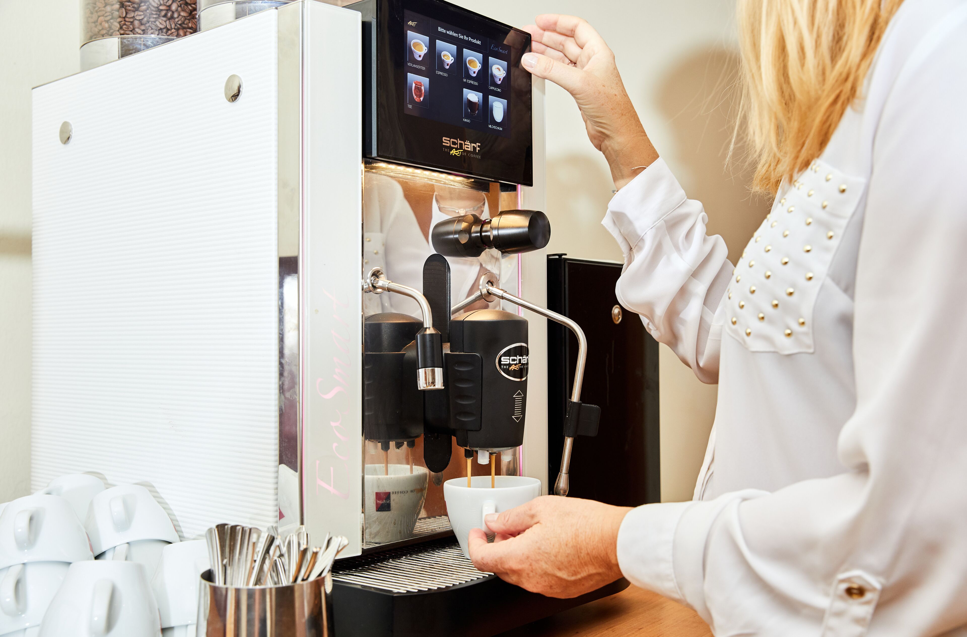 En person betjener en kaffemaskine med berøringsdisplay og holder en kop under dysen.