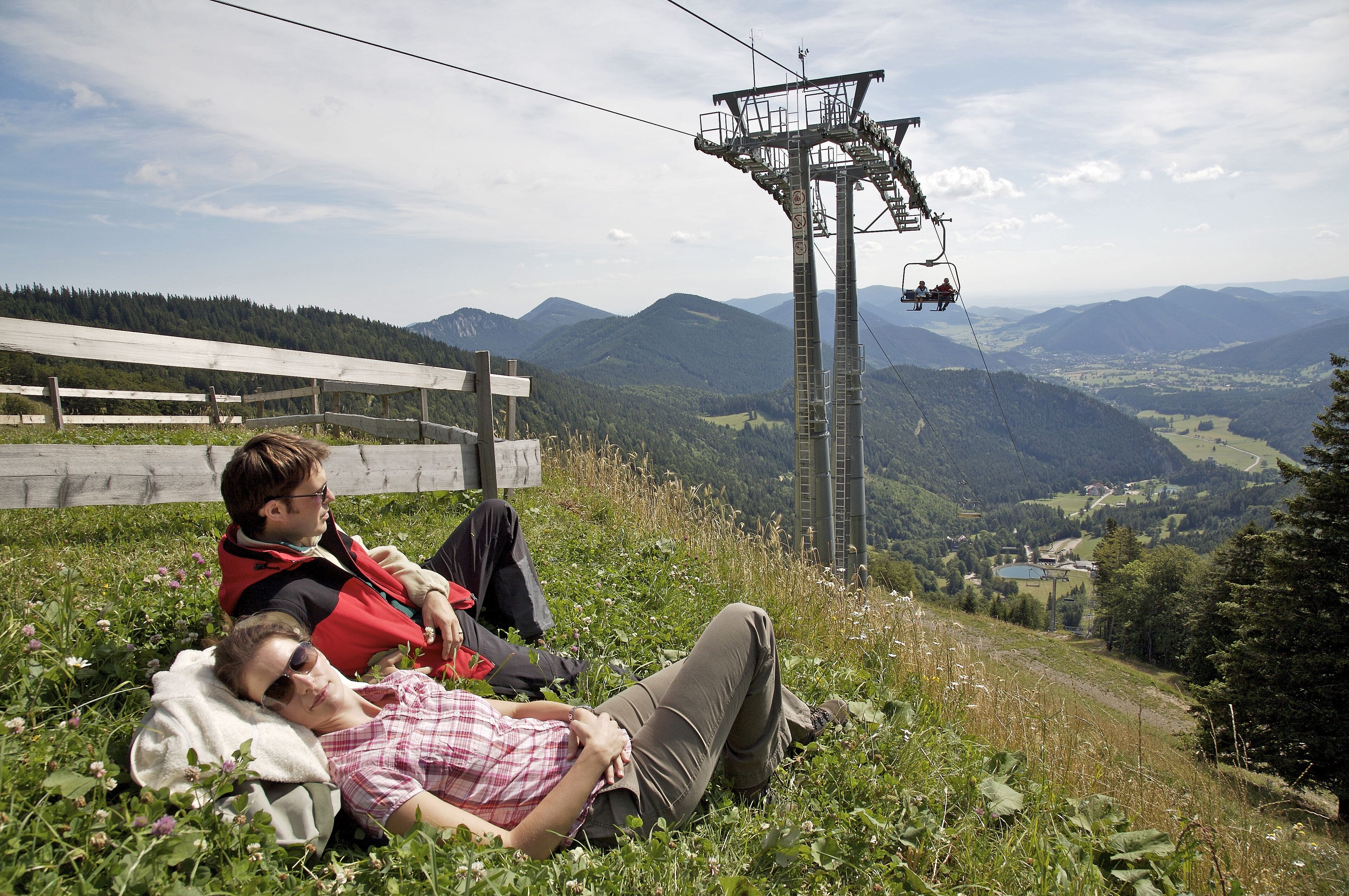 Zwei Personen liegen im Gras, mit Blick auf eine Berglandschaft und einer Seilbahn im Hintergrund.