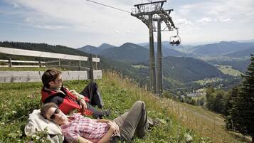 Zwei Personen liegen im Gras, mit Blick auf eine Berglandschaft und einer Seilbahn im Hintergrund.