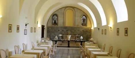 Un comedor con mesas y sillas dispuestas en filas, techos abovedados y una pared de piedra con un ventanal.