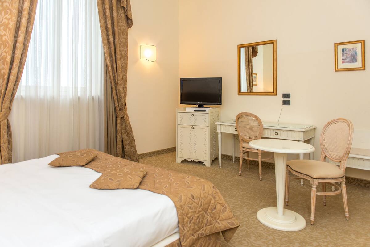 A hotel room with a bed, a desk with a television, two chairs, a small table, and a mirror on the wall.