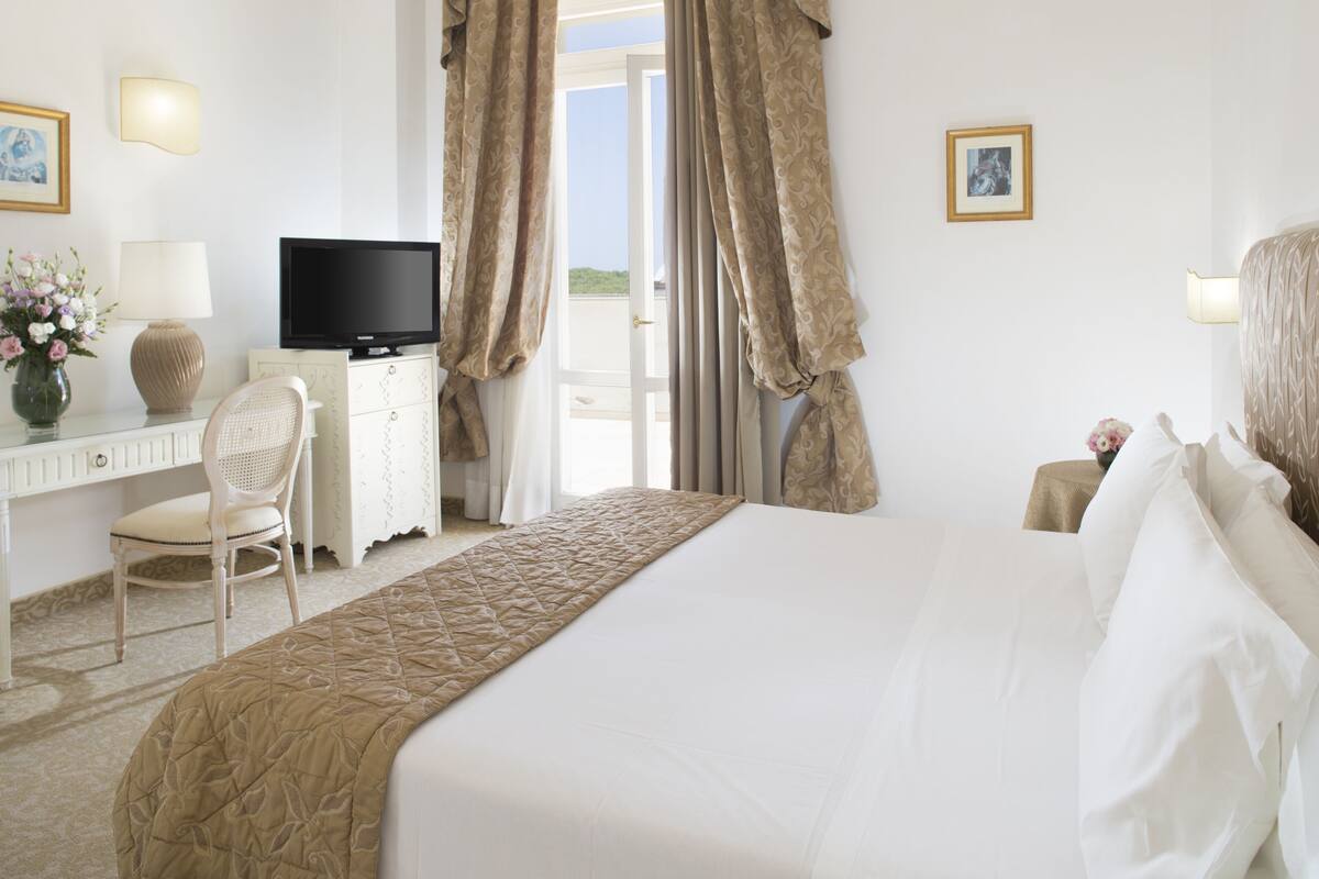 A hotel room with a bed, a desk with a television, a chair, and a window with curtains.