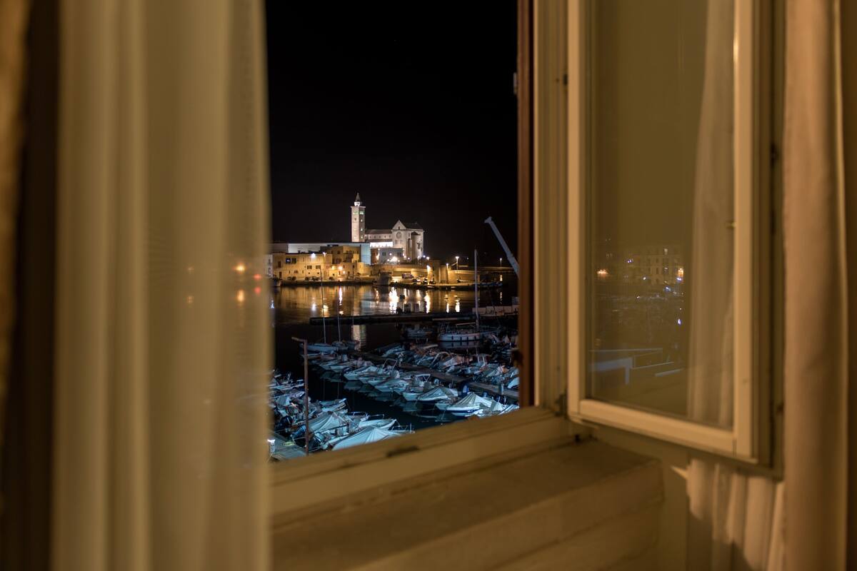 A view through an open window of a marina at night, with boats docked and a building with a tower in the background.