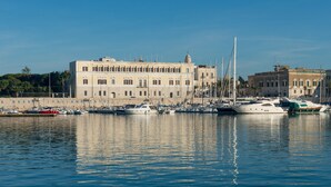 A marina with multiple boats docked, a large building with a classical architectural style, and calm water reflecting the surroundings. - Hotel San Paolo al Convento (Trani)