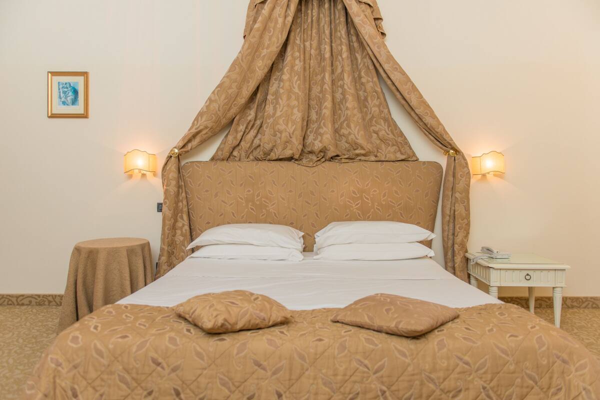 A neatly made bed with a canopy, two bedside lamps, a small round table, and a nightstand with a phone.