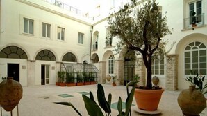 A courtyard with a large tree, potted plants, and a building with arched windows and a balcony. - Hotel San Paolo al Convento (Trani)