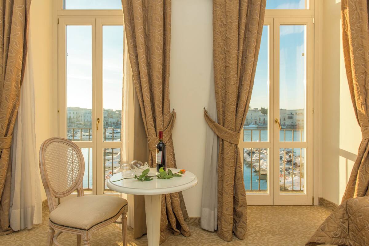 A room with a view of a body of water, a table with a bottle and glasses, a chair, and curtains.