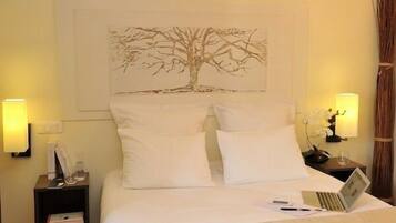 Una cama bien tendida con sábanas blancas, un ordenador portátil sobre la cama y un cuadro colgado en la pared con un árbol.