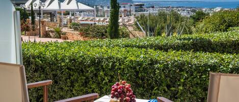 A balcony with a table set with fruit, overlooking a coastal town with buildings and a mountain in the background.