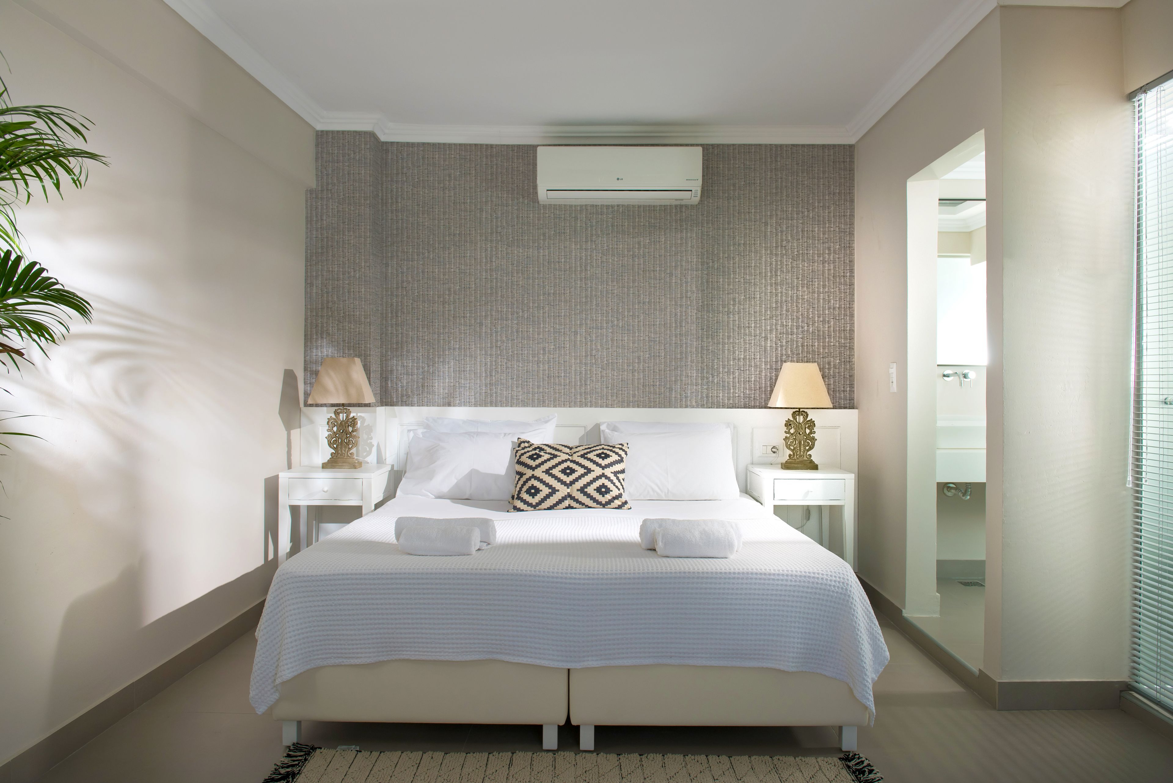 A modern bedroom with a large bed, two bedside tables with lamps, a wall-mounted air conditioner, and a view of a bathroom through an open door.