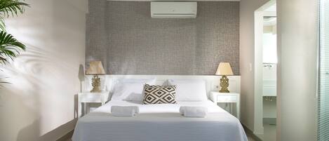 A modern bedroom with a large bed, two bedside tables with lamps, a wall-mounted air conditioner, and a view of a bathroom through an open door.
