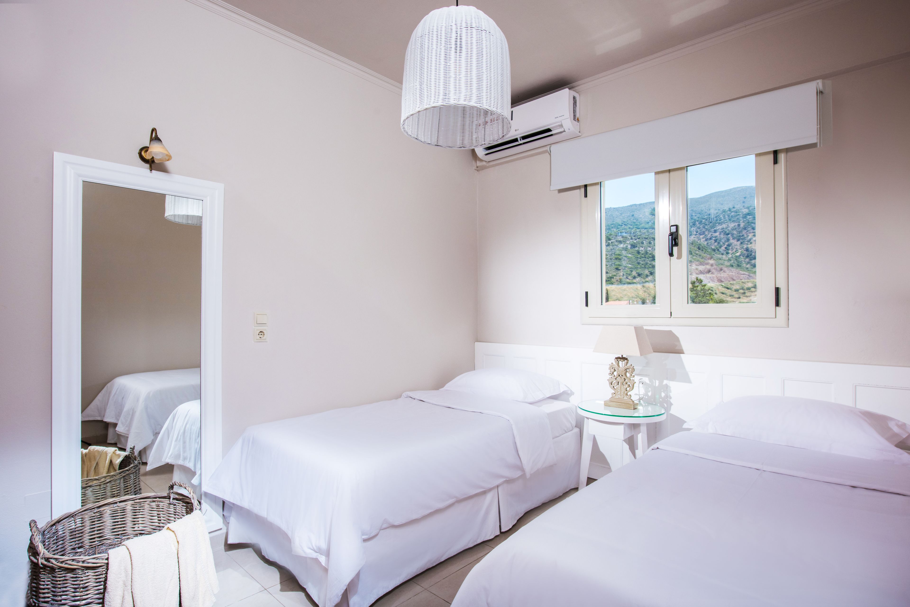 Habitación de hotel con dos camas individuales, una mesita redonda con una lámpara, una cesta de mimbre y vistas al exterior por una ventana.