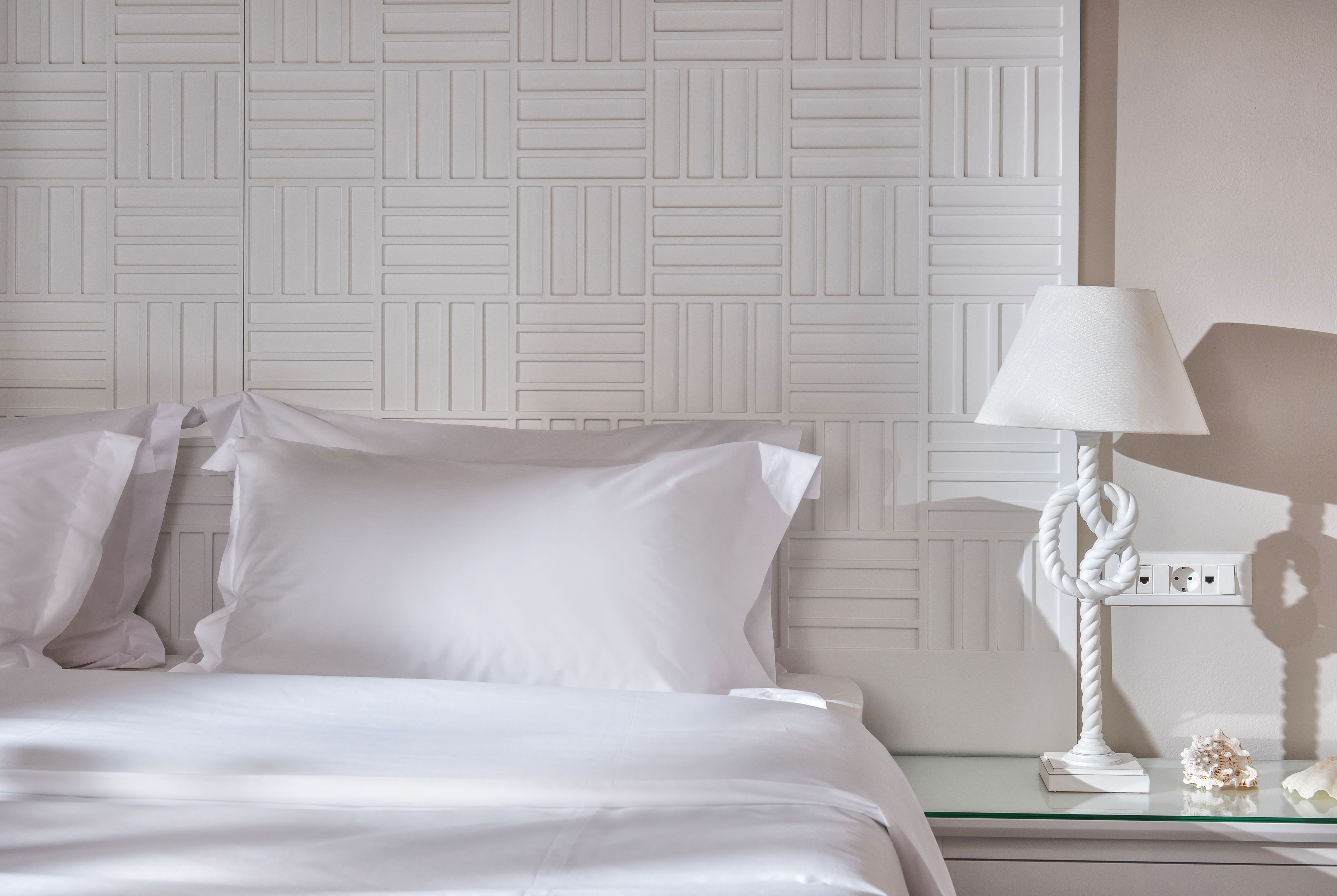A neatly made bed with white linens and pillows, a white textured wall, and a bedside table with a white lamp and decorative item.
