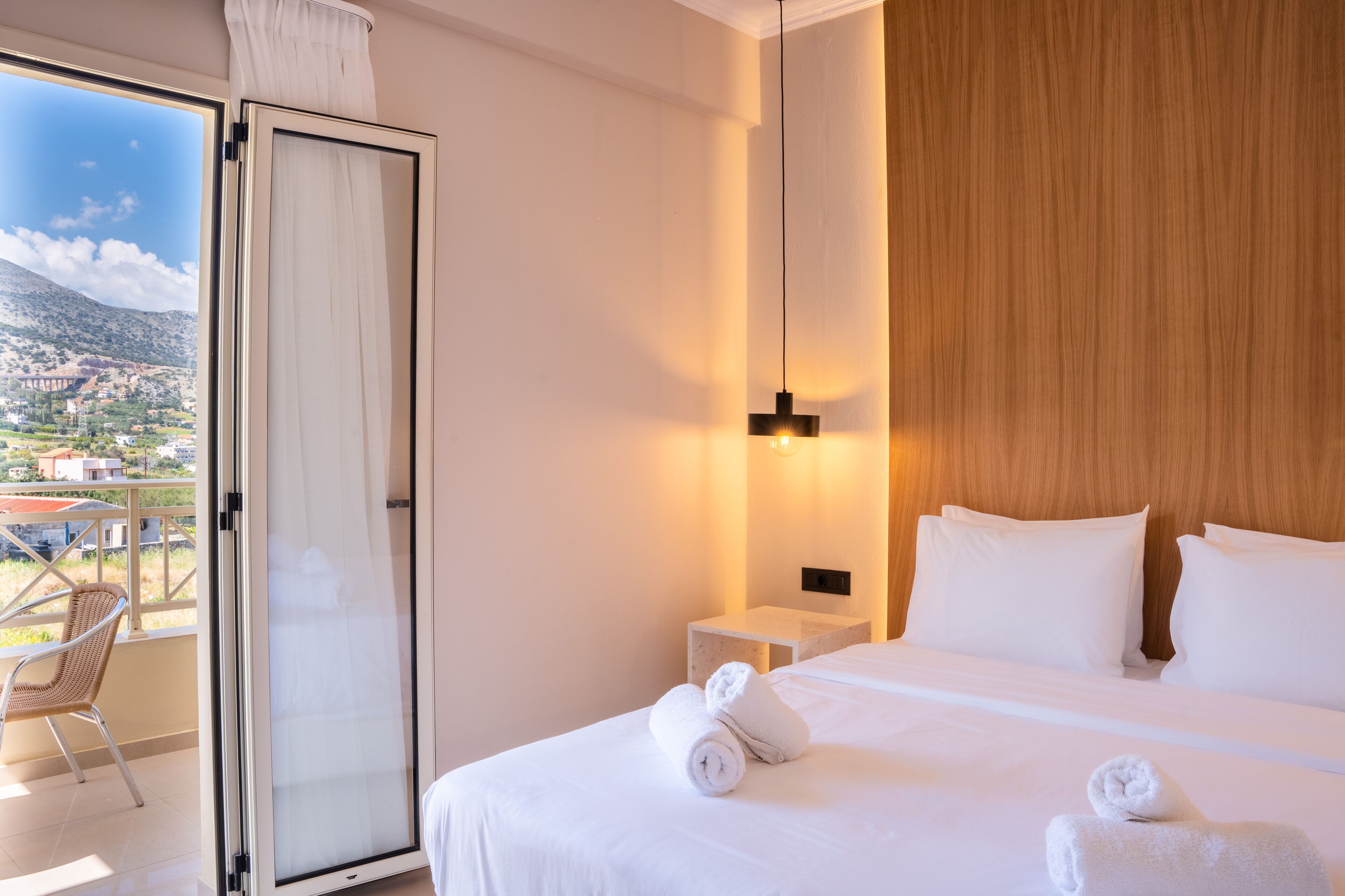 A hotel room with a bed, white linens, a nightstand, a lamp, a window with a view of a mountain and buildings, and a chair by the window.