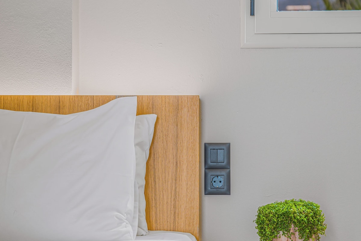 A neatly made bed with white linens, a wooden headboard, a small bedside table with a potted plant, and a wall socket.