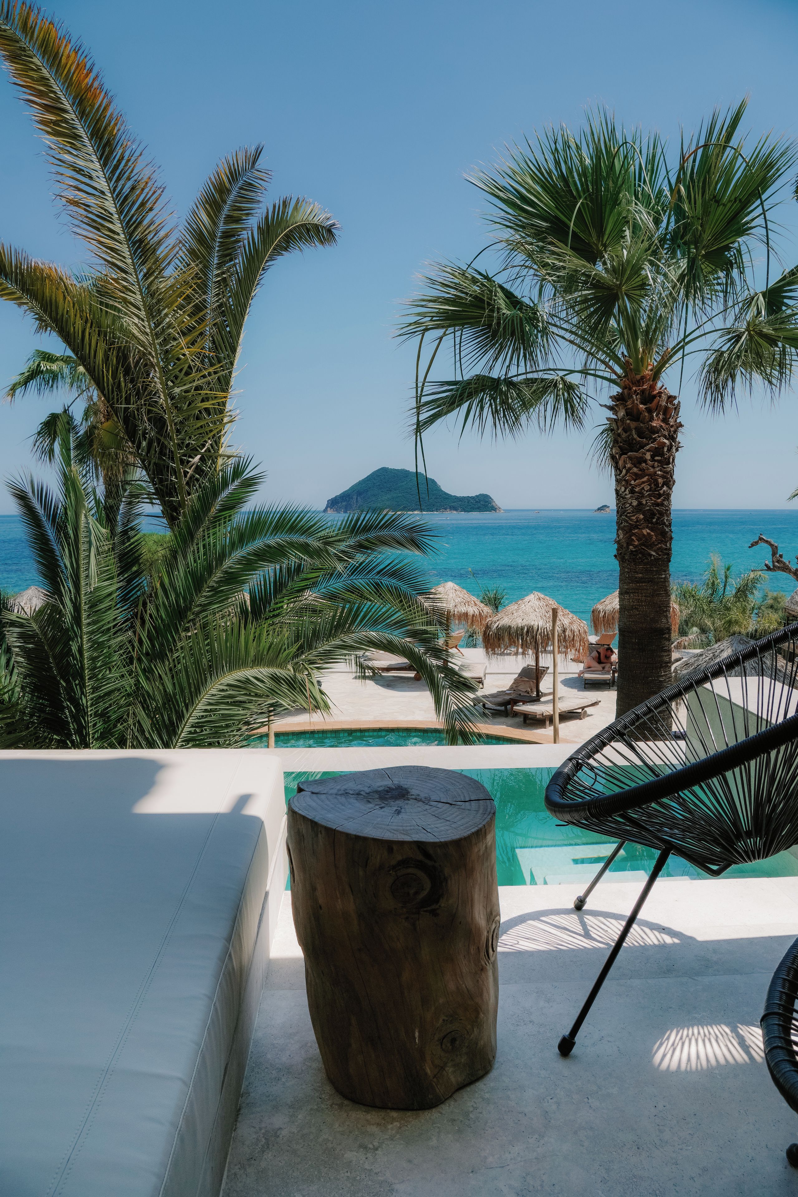Un espace aménagé au bord de la piscine, avec des chaises longues, un tronc d’arbre servant de table, et une vue sur la mer et une île lointaine.