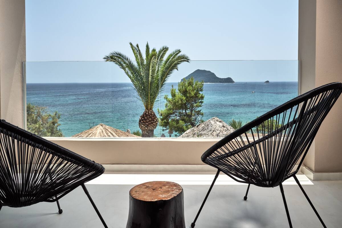 Ein Balkon mit zwei schwarzen Korbstühlen, einem hölzernen Hocker und Blick auf das Meer sowie eine entfernte Insel.