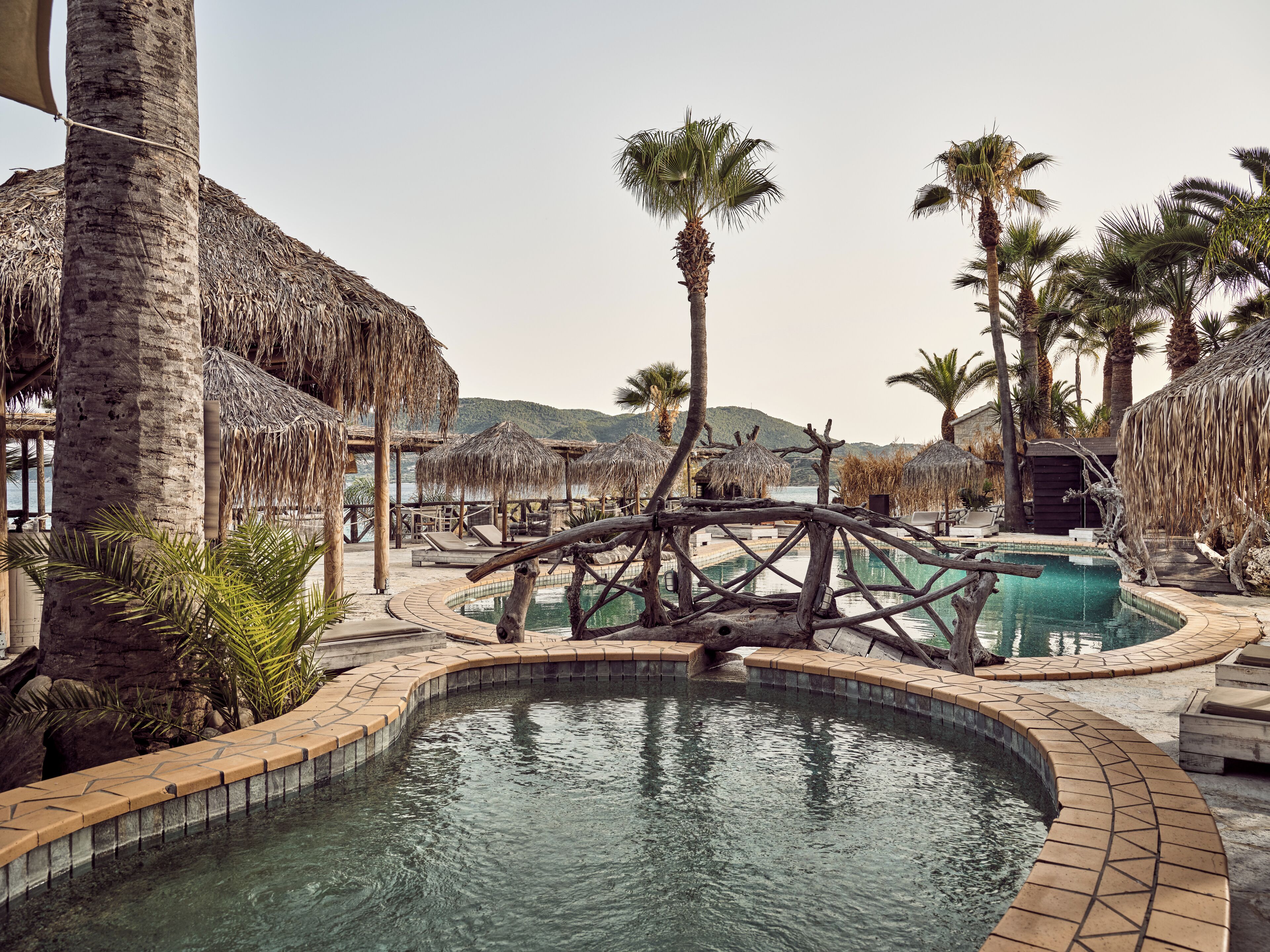 Un área de piscina con una fuente de agua circular, palmeras y sombrillas de paja.