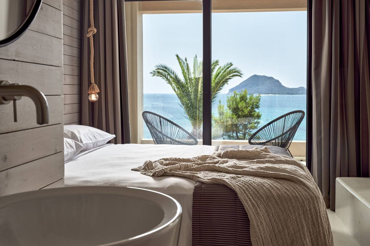 Ein Schlafzimmer mit großem Fenster, einem Bett, einem Badezimmer mit Badewanne und Blick auf das Meer und einen Berg.