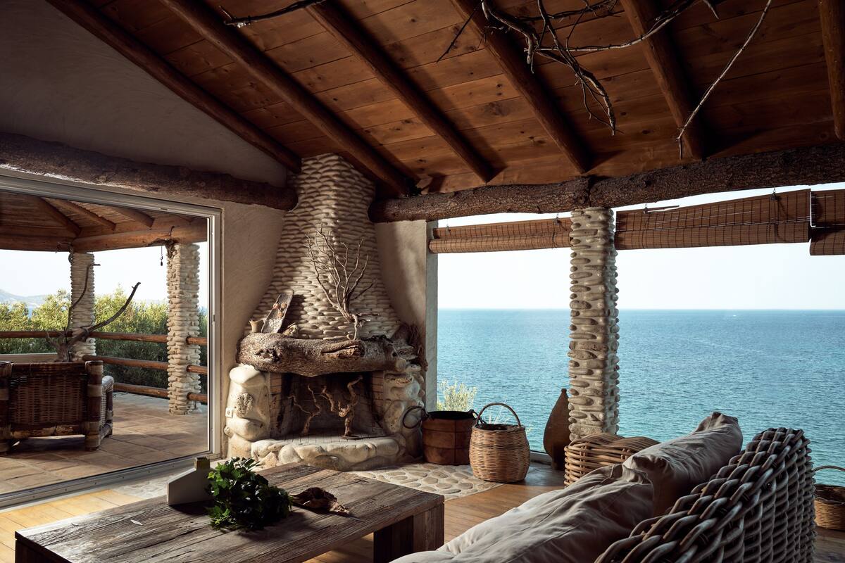 Ein rustikales Interieur mit Kamin aus Stein, Korbmöbeln und Blick auf das Meer.