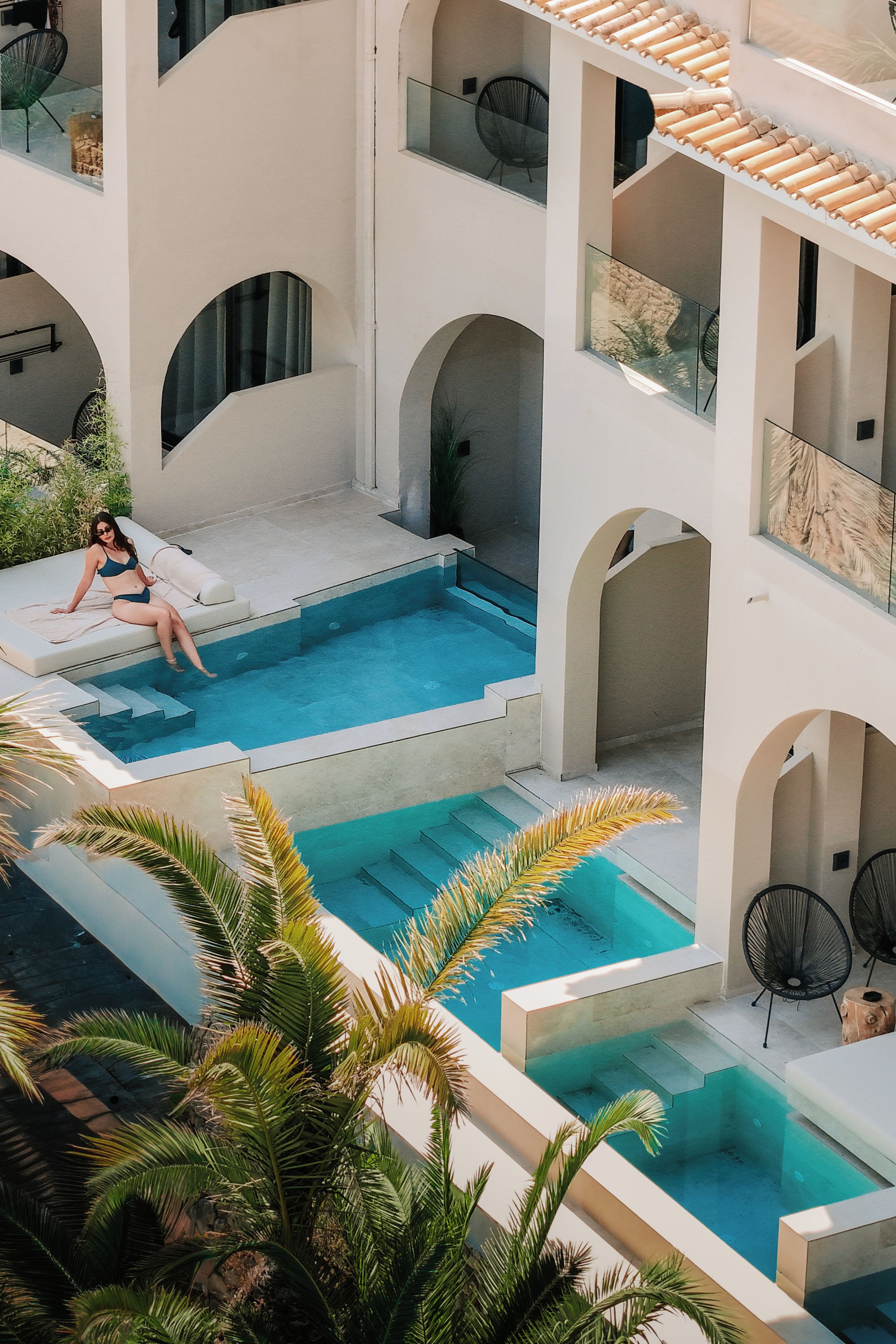 A multi-level swimming pool with clear blue water, surrounded by white walls and arches, and palm trees in the foreground.