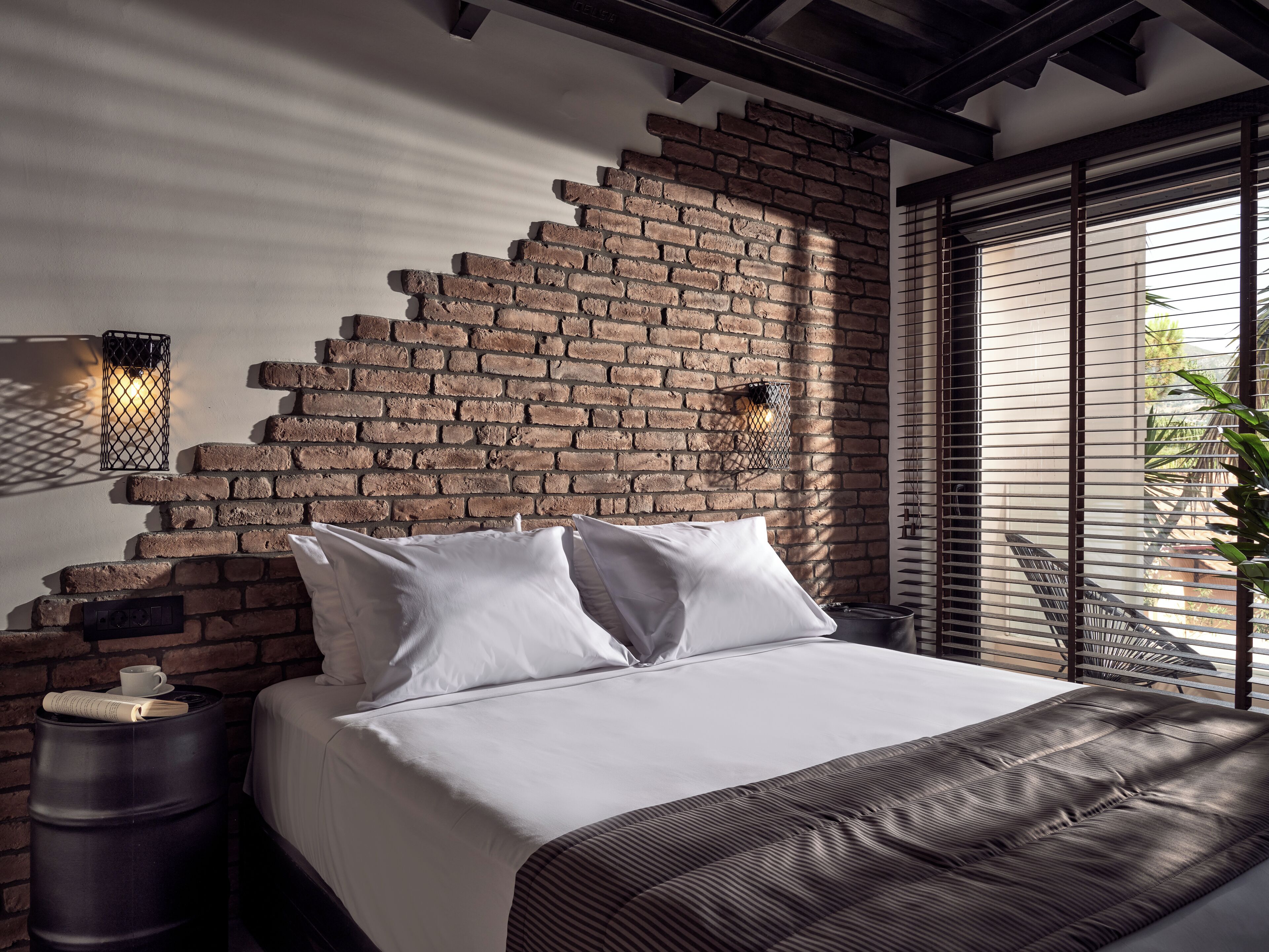 A bedroom with a brick wall, a bed with white bedding, two bedside tables, and a window with blinds.