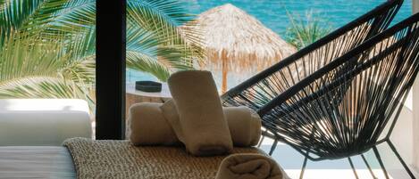 A room with a view of the sea, a bed, a chair, and a thatched umbrella.