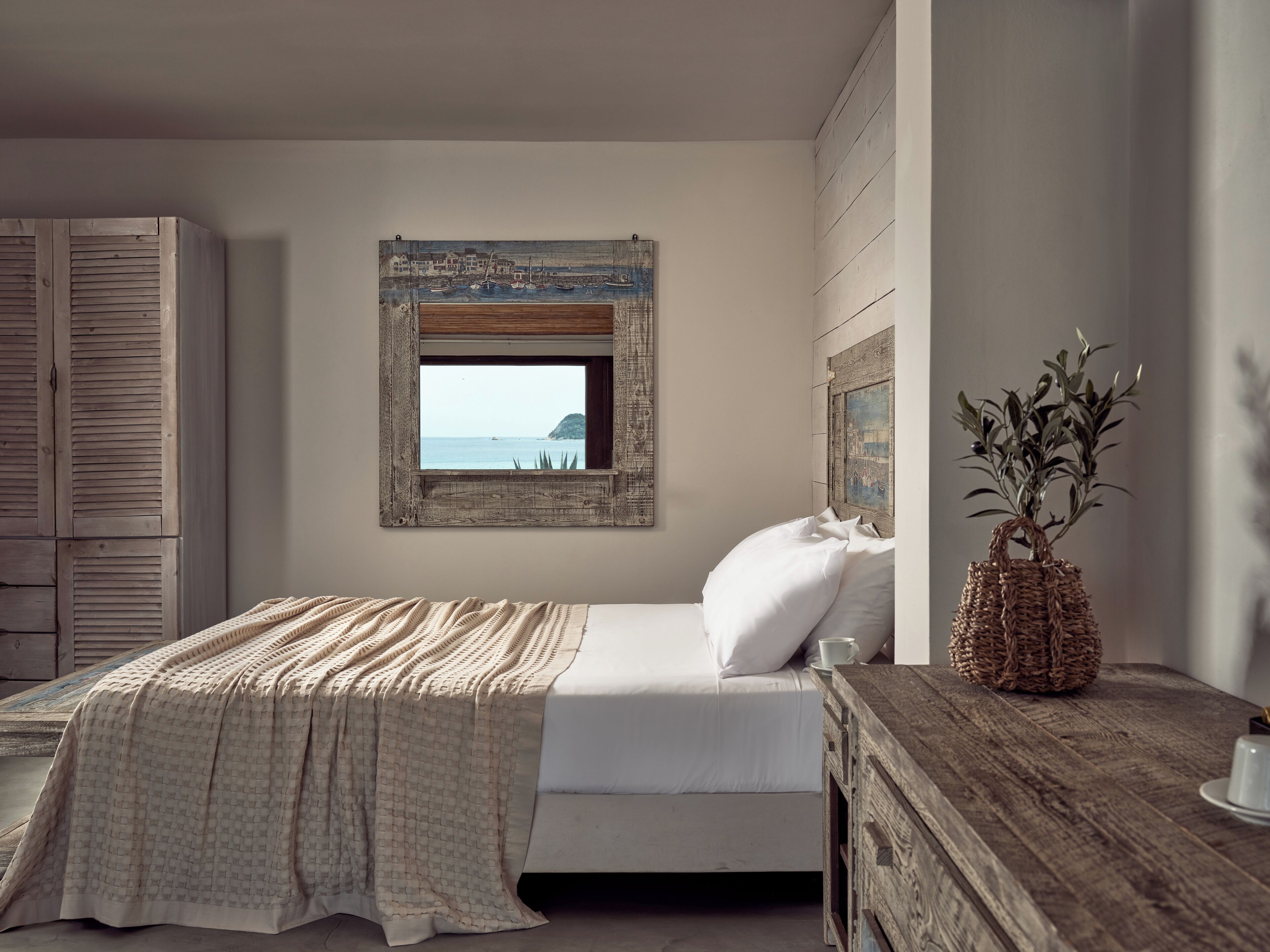 A bedroom with a bed, a wooden dresser, a window with a view of the sea, and a potted plant.