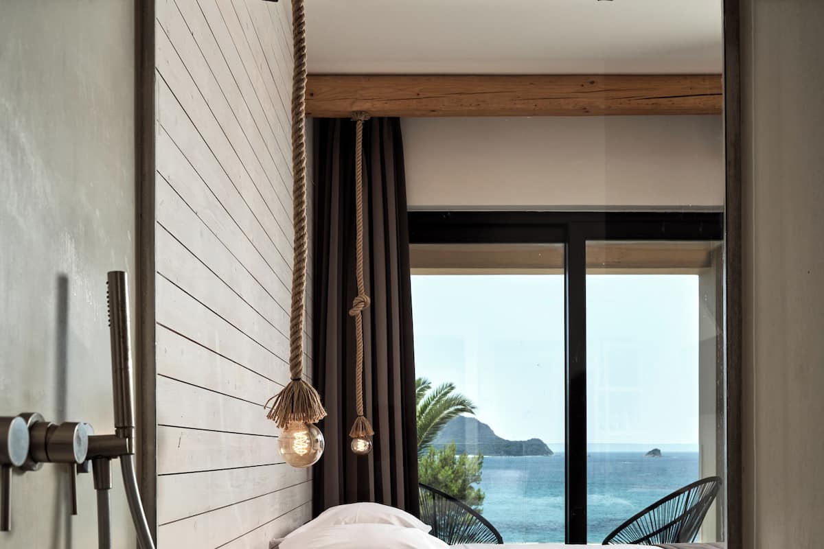 A modern bedroom with a wooden slatted wall, a bed with white bedding, a bedside table with a cup and book, a hanging light fixture, and a view of the sea through large windows.
