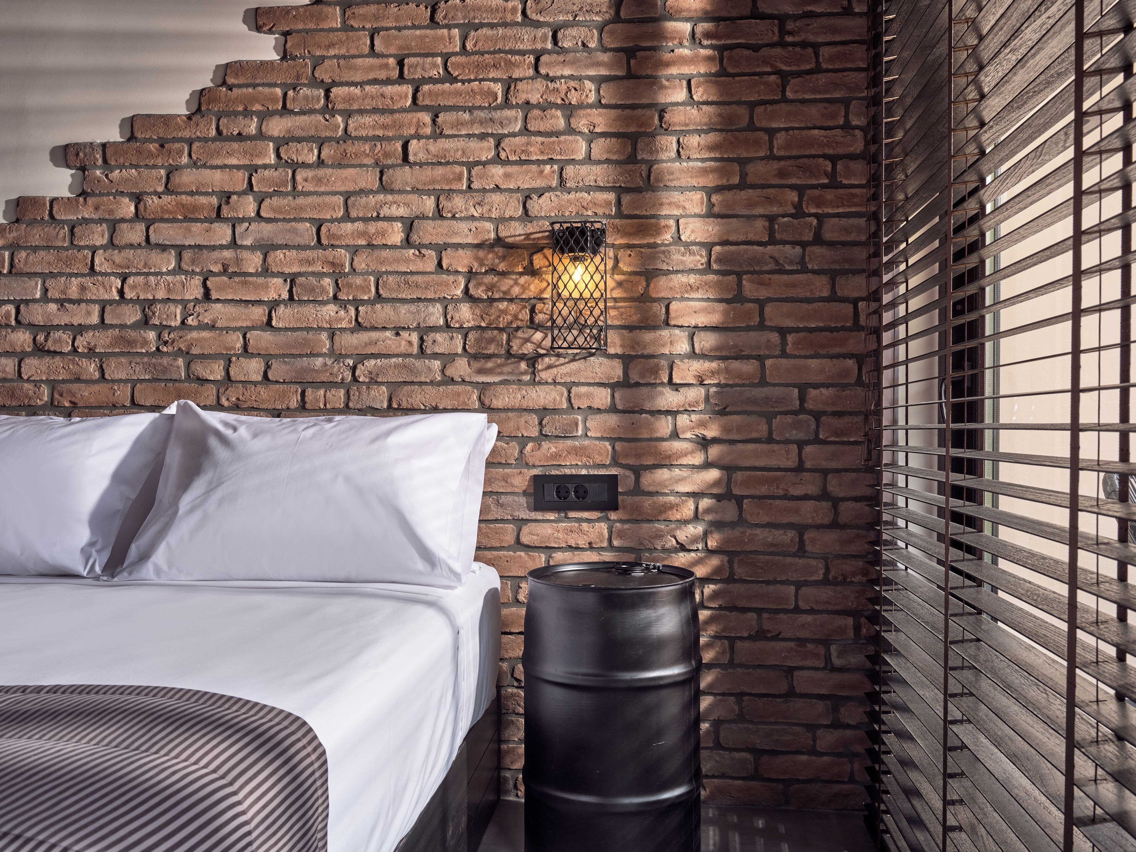 Un dormitorio con pared de ladrillo, cama con sábanas blancas, un barril negro y una ventana con persianas.