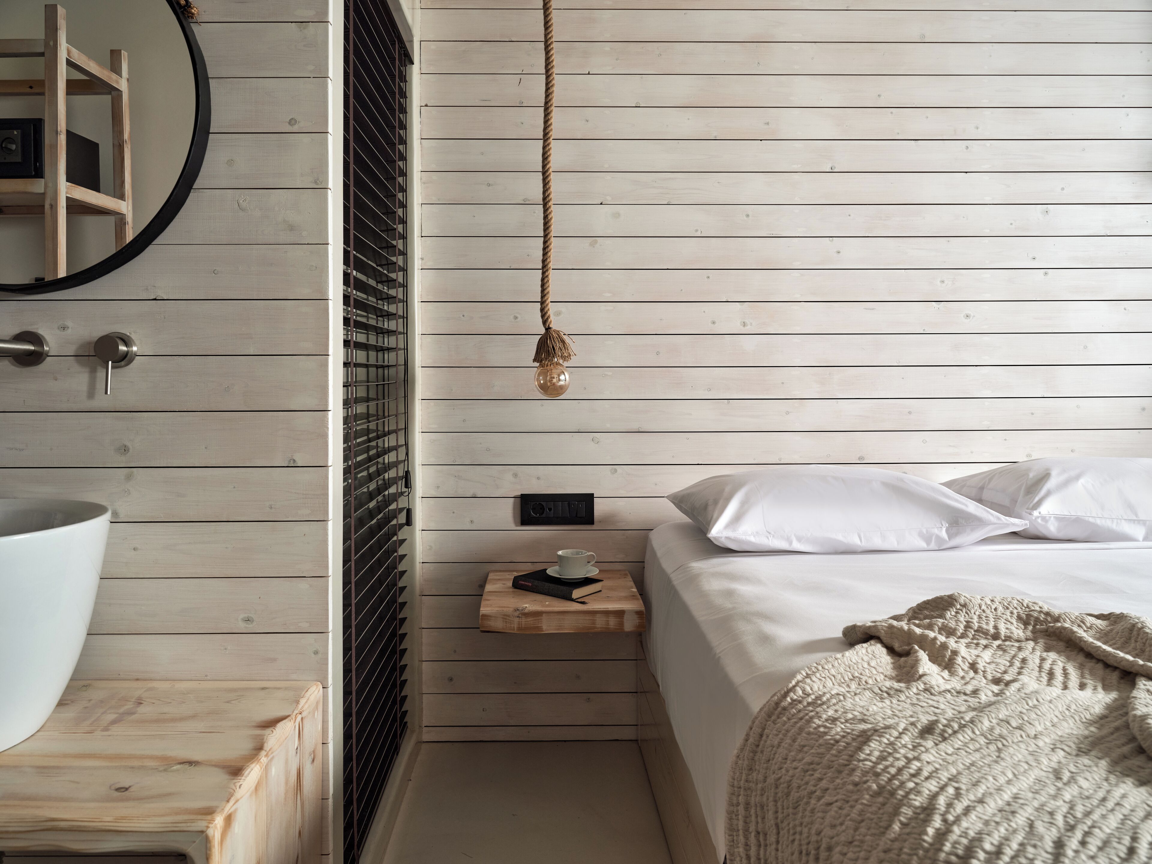 A bedroom with a wooden slat wall, a round mirror, a bedside table with a cup and a book, a bed with white linens, and a hanging light fixture.