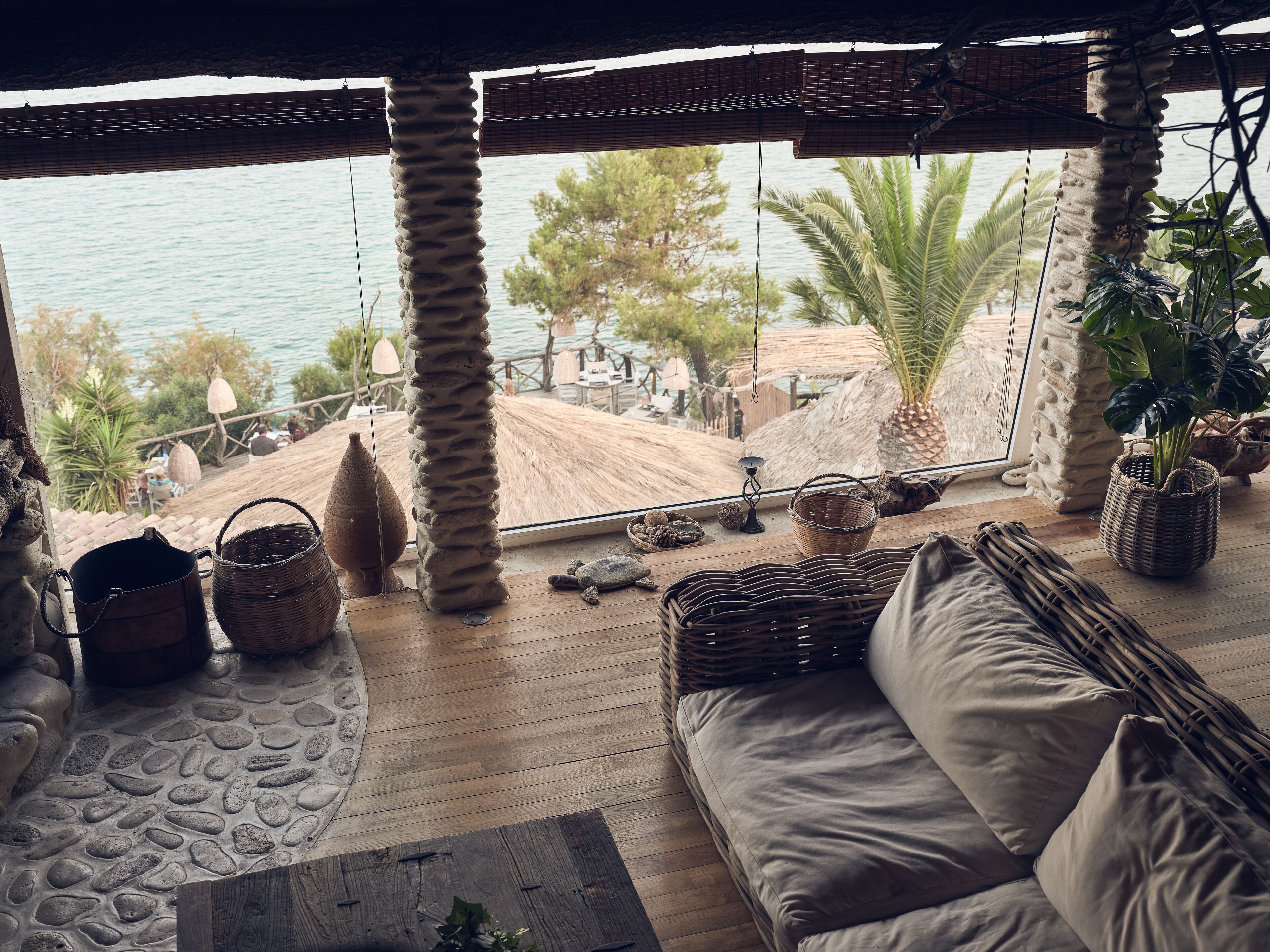 A wooden deck with a sofa and cushions, a stone floor, and a view of the sea and palm trees.
