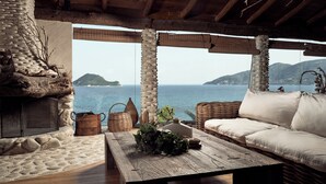 A rustic interior with a stone fireplace, a wooden table with a plant, a wicker sofa, and a view of the sea and mountains. - Gloria Maris Hotel Suites & Villas (Zakynthos)