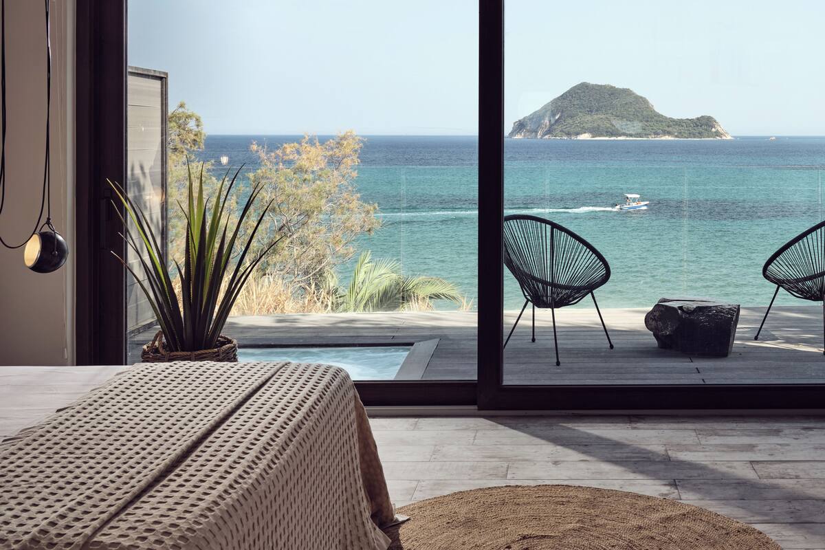 Ein Schlafzimmer mit großem Fenster mit Blick auf einen Pool und das Meer, ausgestattet mit einem Bett, zwei Stühlen und einer Topfpflanze.