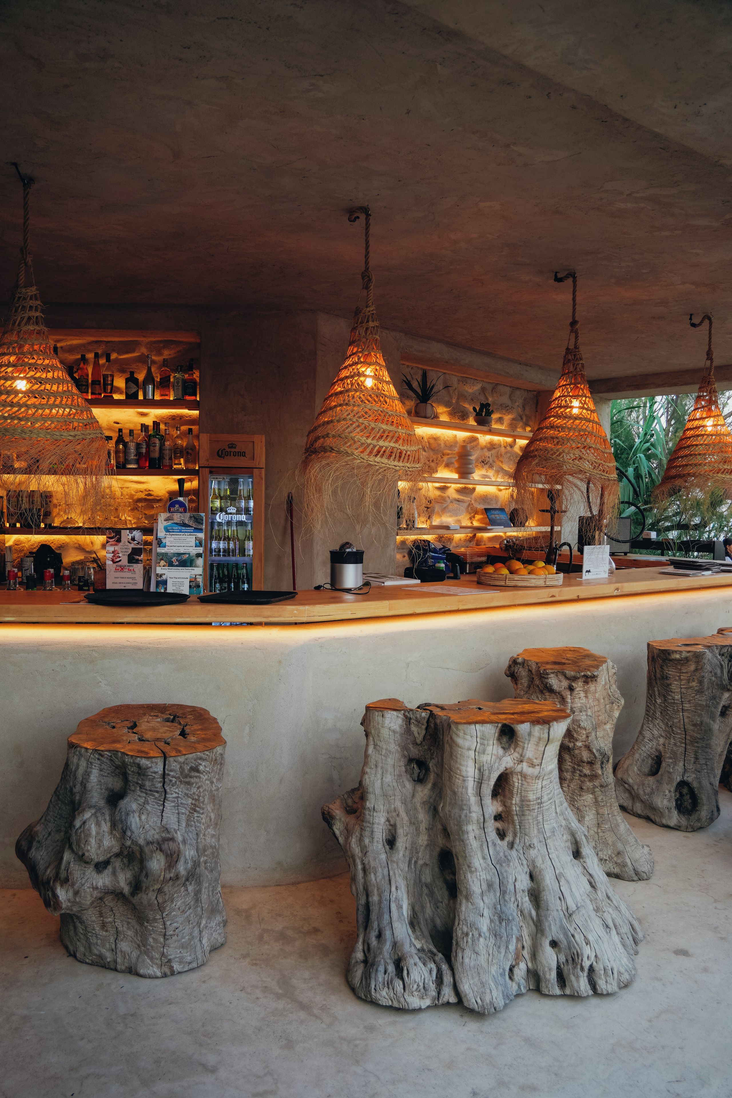 A bar area with wooden stools, a Corona refrigerator, and hanging woven light fixtures.