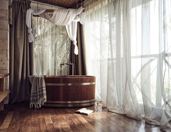 Une baignoire en bois dans une pièce au plancher en bois, une fenêtre ornée de rideaux légers et un banc en bois rustique.