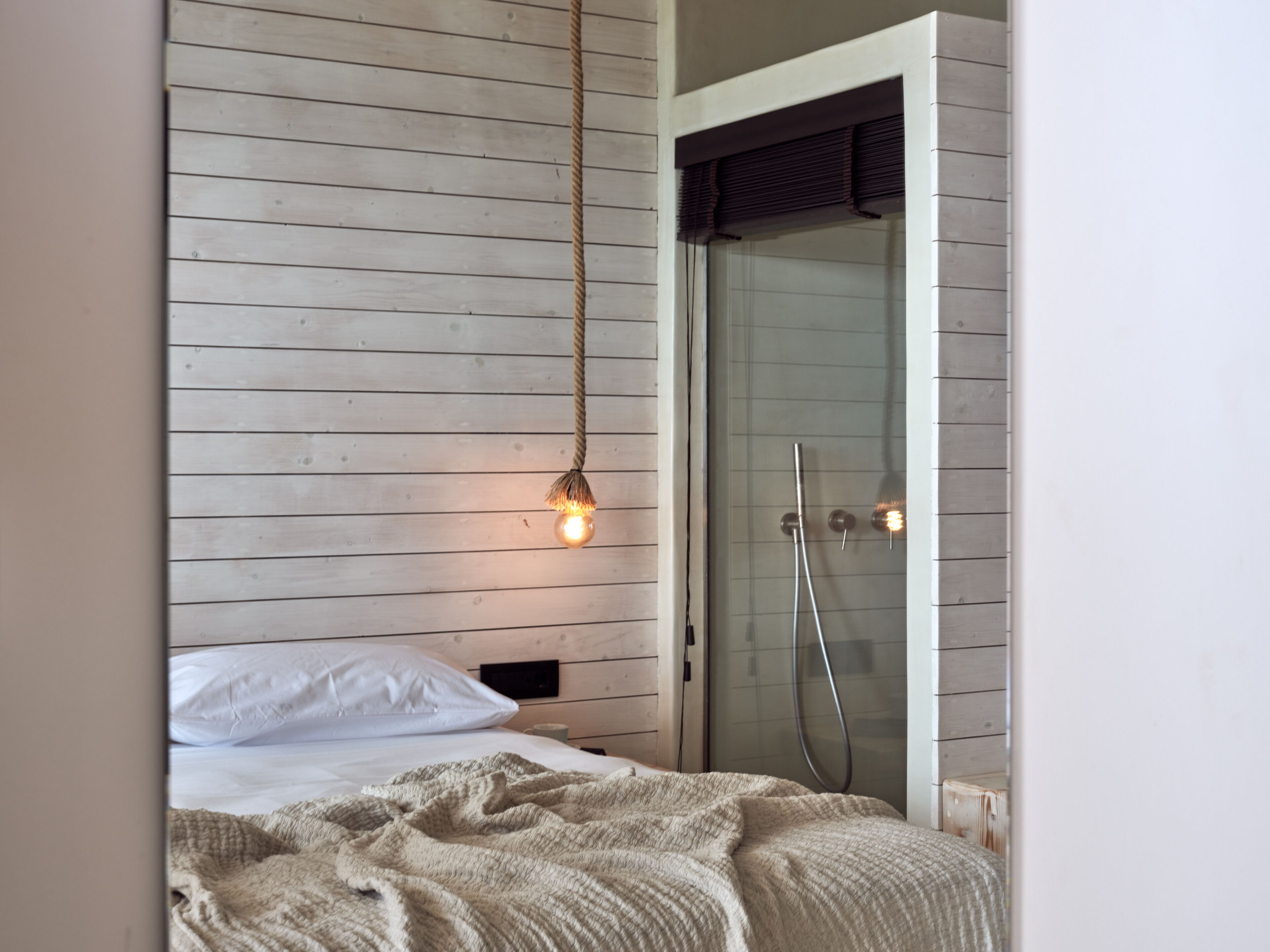 Camera da letto con parete in legno a listelli, lampadario a sospensione, letto con biancheria bianca e zona doccia con porte in vetro.