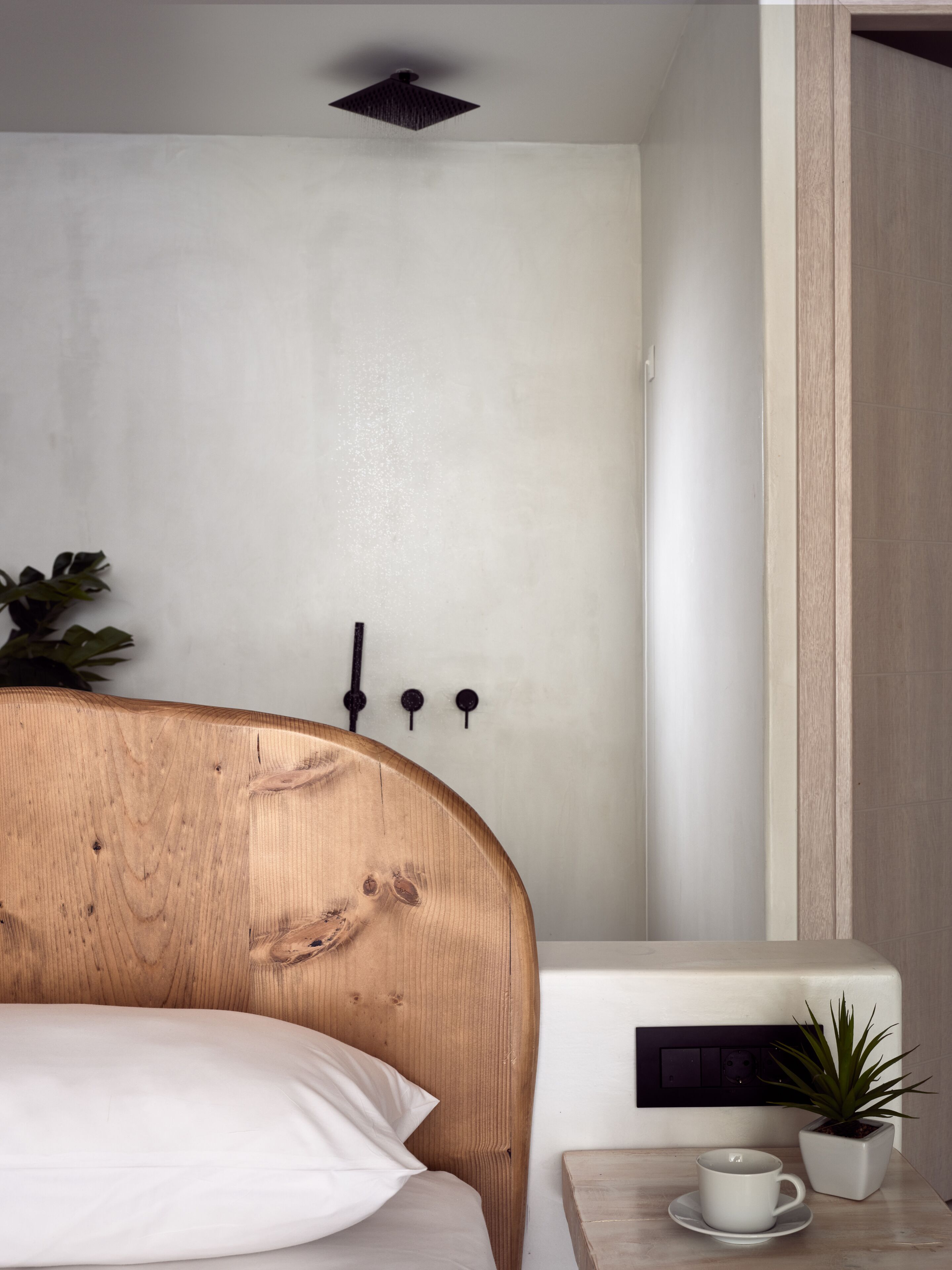A bedroom with a wooden headboard, a white pillow, a bedside table with a cup and a potted plant, and a wall-mounted light fixture.