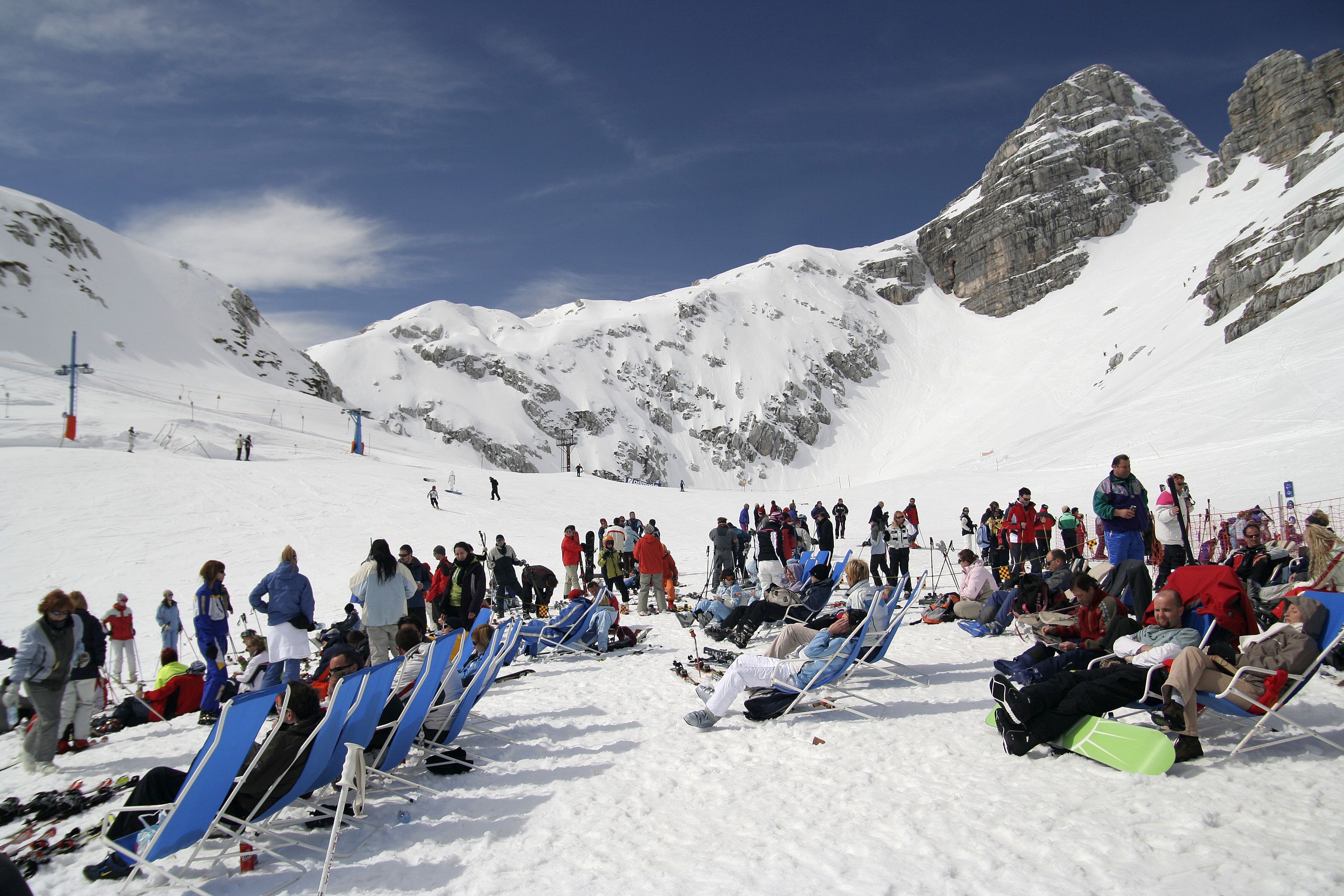 A snow-covered ski slope with people sitting on blue chairs, likely taking a break.