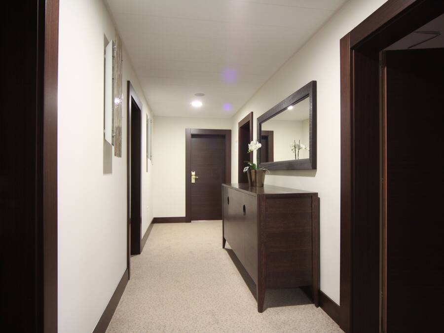 A hallway with a wooden door at the end, a dark wooden dresser with a mirror and a potted plant, and a light-colored carpet.