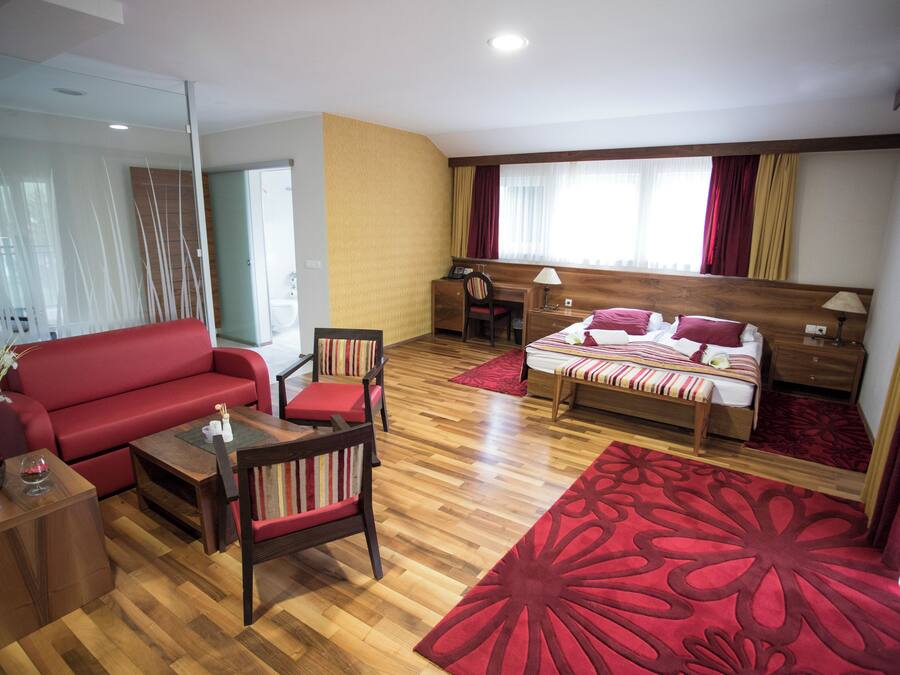 A hotel room with a wooden floor, a red sofa, a desk with a chair, a bed with a red and white striped bedspread, and a large red floral rug.