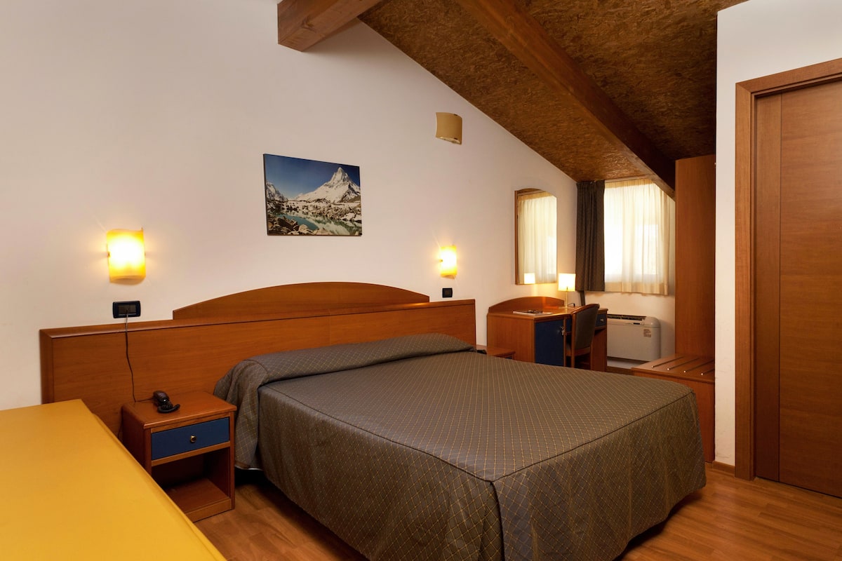 A hotel room with a wooden bed, bedside tables, a desk with a chair, a mirror, and a window with curtains.