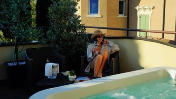A person relaxing by a hot tub on a balcony with a view of a building and a tree.