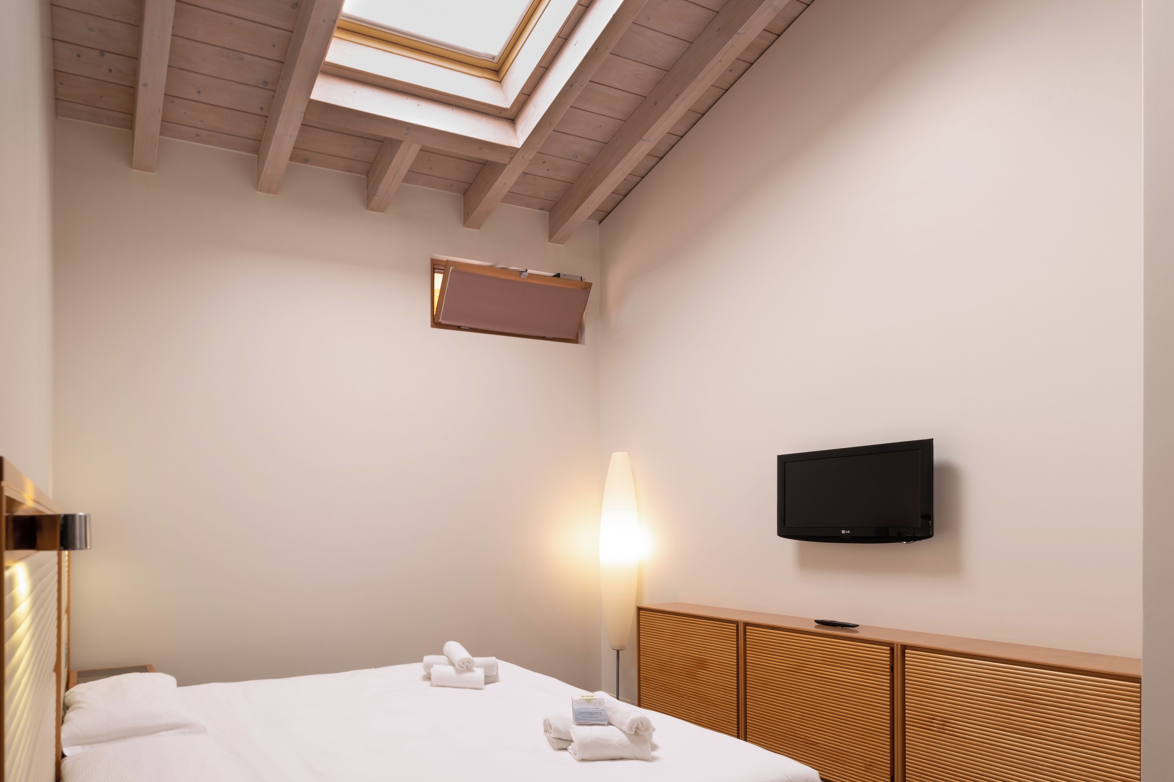 Habitación de hotel con cama, televisión de pantalla plana, lámpara de noche y tragaluz.