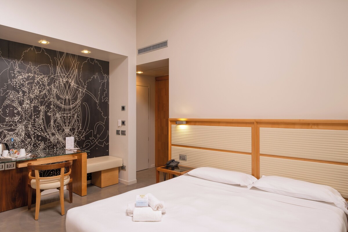 Une chambre d’hôtel comprenant un lit, un bureau avec une chaise, une grande œuvre d’art abstraite accrochée au mur, une tête de lit en bois et une table de chevet avec un téléphone et une lampe.